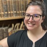 Emily Stensloff, a white woman with brown hair and glasses, standing in front of a bookcase full of 19th century books.